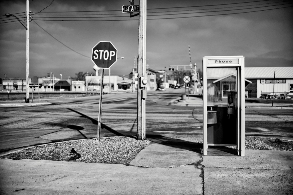 A black and white photo of a stop sign and phone booth on a street corner in a small town. 